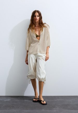 Femme aux longs cheveux ondulés porte un cardigan en tricot beige par-dessus un haut olive, un pantalon blanc ample court avec des liens, et des tongs noires, debout contre un mur blanc.