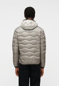 Άνδρας που φαίνεται από πίσω, φορώντας γκρι ανοιχτό χρώμα μπουφάν με κουκούλα και μαύρο παντελόνι, σε απλό φόντο.