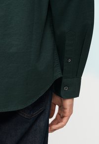 Κοντινό πλάνο από το μπράτσο ενός ατόμου που φοράει μανσέτα πουκάμισου σκούρου πράσινου χρώματος με κουμπιά, με το χέρι του να ακουμπά κοντά στην πίσω τσέπη σκούρων μπλε τζιν.