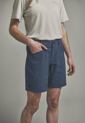 Person trägt beige T-Shirt und marineblaue Shorts, eine Hand in der Tasche, zeigt Vitiligo an Armen und Beinen.