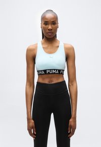 Ευέλικτη γυναίκα με αλογοουρά που φοράει ανοιχτό μπλε αθλητικό σουτιέν Puma και ψηλόμεσο μαύρο κολάν, στέκεται μπροστά σε απλό ανοιχτό φόντο.