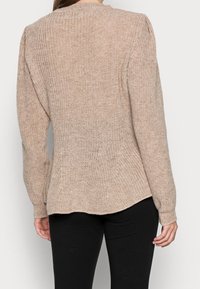 Beżowy sweter dziany z długimi rękawami i okrągłym dekoltem. Teksturowany, luźny fason z delikatnie zaokrąglonym dołem, w zestawie z czarnymi spodniami.
