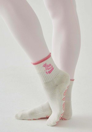 Stopy w bladoróżowych rajstopach, ubrane w kremowe skarpetki z różową lamówką i sercowatymi antypoślizgowymi kropkami na podeszwach, stojące na palcach.