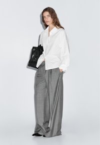 Weißes Hemd mit Kragen, kombiniert mit grauen, gestreiften, weit geschnittenen Hosen. Das Modell hält eine schwarze Ledertasche. Glatte Texturen und locker sitzendes Design.