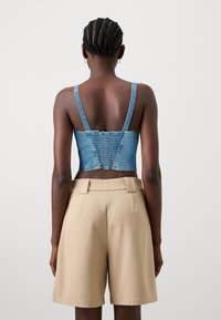 Top cropped in denim blu con retro arricciato e spalline regolabili, abbinato a un pantalone corto beige sartoriale, mostrato da dietro.
