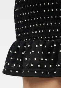 Μαύρη φούστα με λευκές πουά, με υφασμένη πάνω περιοχή με σφηκοφωλιά και βολάν στο τελείωμα πάνω από το γόνατο.