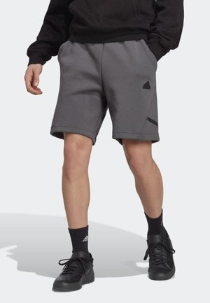 Mężczyzna w ciemnoszarych szortach Adidas, czarnych skarpetach Adidas, czarnych sneakersach i czarnej bluzie, stojący z rękami w kieszeniach.