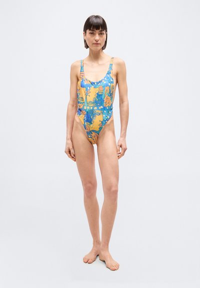 Γυναίκα που στέκεται ξυπόλυτη φορώντας πολύχρωμο ολόσωμο μαγιό με αφηρημένα μοτίβα προσώπων σε μπλε, πορτοκαλί και κίτρινο χρώμα και αστεριών σε απλό φόντο.