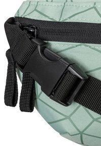 Gros plan d'une boucle en plastique noire et d'une sangle réglable attachées à un sac vert clair avec un motif géométrique et une fermeture éclair noire.