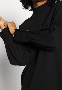 Blusa nera a maniche lunghe con collo alto. Presenta maniche arricciate e un dettaglio con bottone al polsino. Tessuto morbido e testurizzato con una vestibilità rilassata.