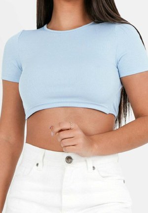 Woman wearing a light blue ribbed crop top and high-waisted white jeans, with long dark hair and a gold ring on her left hand.