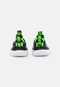 Chaussures de sport noires avec semelles blanches, présentant des accents vert fluo, des languettes de traction et une texture lisse. Logo Nike notable en blanc.