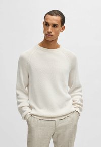 Cremfarbener, gerippter Strickpullover mit Rundhalsausschnitt und Raglanärmeln, kombiniert mit hellbeigen Hosen. Weiche Textur und lässige Passform.