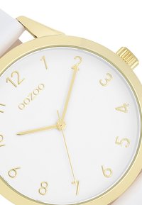 Montre dorée avec un cadran rond blanc, des index horaires minimalistes dorés et des aiguilles dorées fines. Le bracelet blanc complète le design.