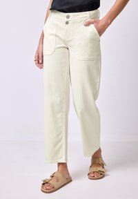 Žena v krémových straight-leg kalhotách s velkými předními kapsami a béžových sandálech se zlatými přezkami, stojící na neutrálním pozadí.