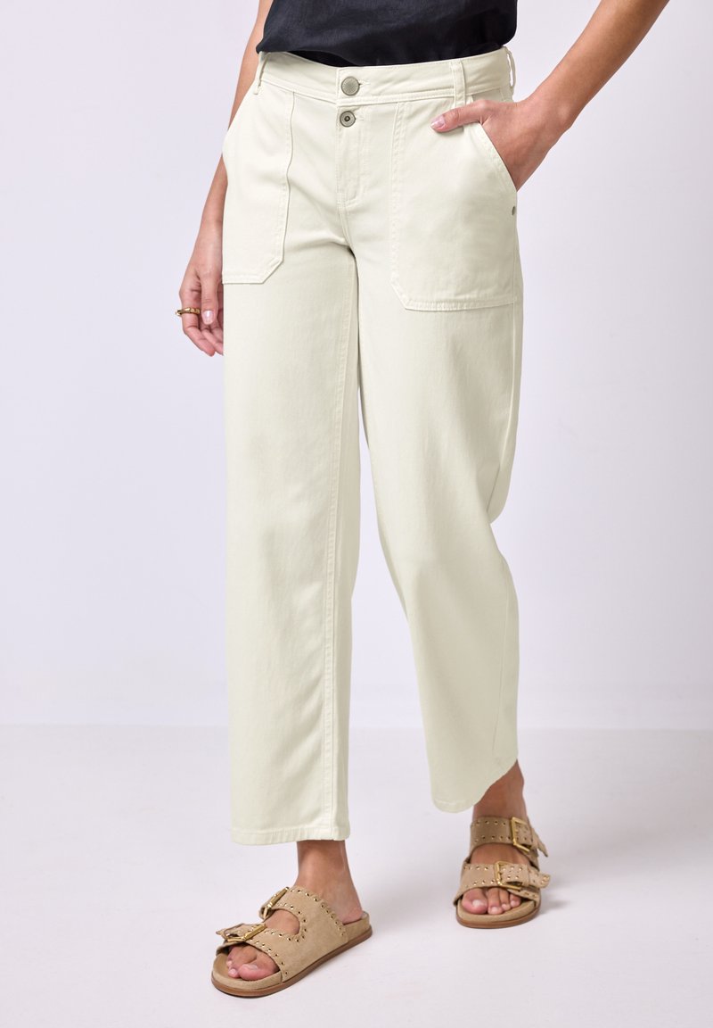 Žena v krémových straight-leg kalhotách s velkými předními kapsami a béžových sandálech se zlatými přezkami, stojící na neutrálním pozadí.