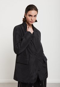 Crni, oversized blejzer od glatke tkanine; ima izrezani ovratnik, dva prednja džepa i zatvaranje na jedan dugme.