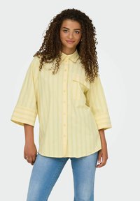 Gulrandig skjorta med krage, korta ärmar och bröstficka, tillverkad av lättviktsmaterial. Matchad med blå jeans.
