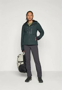 Inimene seisab seljas tumed roheline jope, tumedad püksid, mustad matkajalanõud ning hoiab ühes käes beeži ja musta seljakotti.