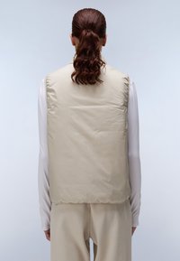 Gilet rembourré beige au toucher lisse, avec un col montant et un ourlet arrondi, porté sur un top blanc à manches longues.