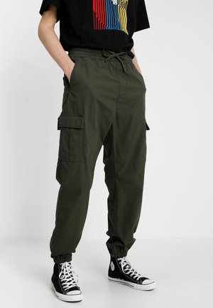 Personne portant un pantalon cargo vert olive avec des poignets élastiques et des baskets montantes noires, debout les mains dans les poches.