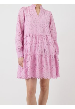 Robe en dentelle ajourée rose clair avec manches longues, jupe à volants, ourlet festonné et encolure en V portée par une personne debout.