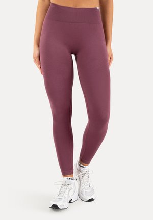 Kvinde iført højttaljede mauve leggings og hvide sneakers med "SMILE" synligt på sokkerne, stående mod en ensfarvet baggrund.