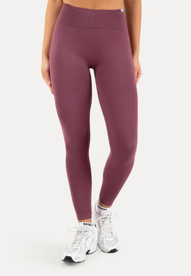 Femme portant des leggings taille haute mauve et des baskets blanches avec le mot "SMILE" visible sur les chaussettes, debout devant un fond uni.