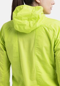 Leichte, leuchtend limegrüne Jacke mit Kapuze. Sie besteht aus strukturiertem Stoff mit Mesh-Details und Nähten entlang des Rückens für eine bessere Luftzirkulation.