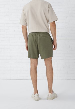 Homme portant une chemise beige à manches courtes, un short vert olive et des baskets blanches, debout face à un mur en briques blanches.