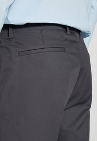 Pantalon gris foncé avec une texture lisse et une poche arrière. Associé à une chemise bleu clair rentrée à la taille.