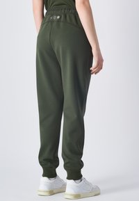 Donkergroene sweats met een gebogen tailleband, elastische manchetten en drie decoratieve knopen op de achterkant. Gepaard met witte sneakers.