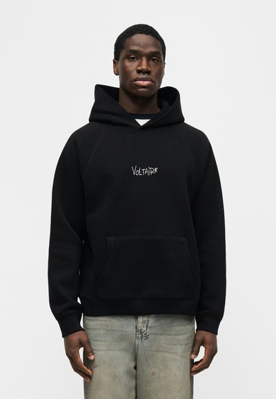 Νεαρός άνδρας που φοράει μαύρο φούτερ με κουκούλα και το κείμενο "Voltaire" στο μπροστινό μέρος και ανοιχτό γκρι τζιν, στέκεται μπροστά σε απλό λευκό φόντο.