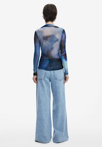 Transparent long-sleeve top with abstract blue and black patterns, paired with wide-leg light blue denim jeans featuring patch pockets.