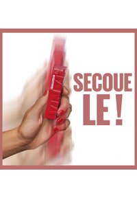 Un emballage rectangulaire rouge tenu dans une main, présentant une surface texturée et un texte en gras. L'arrière-plan est flou, mettant en valeur le produit.