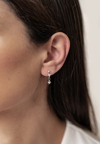 Srebrne kolczyki w kształcie kropli z małymi, przezroczystymi kryształkami, charakteryzujące się delikatnym wzorem łańcuszka i subtelnym blaskiem, zawieszone na przekłutym uchu.