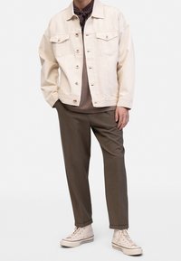 Roomkleurig spijkerjack met knoopsluiting en twee voorzakken, gedragen over een bruine trui. Olijfgroene broek en beige sneakers maken de outfit compleet.