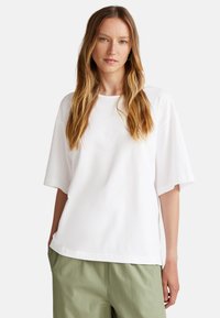 Bílý oversized tričko z jemné látky s kulatým výstřihem a krátkými rukávy, spárované se světle zelenými kalhotami. Jednoduchý, čistý design.