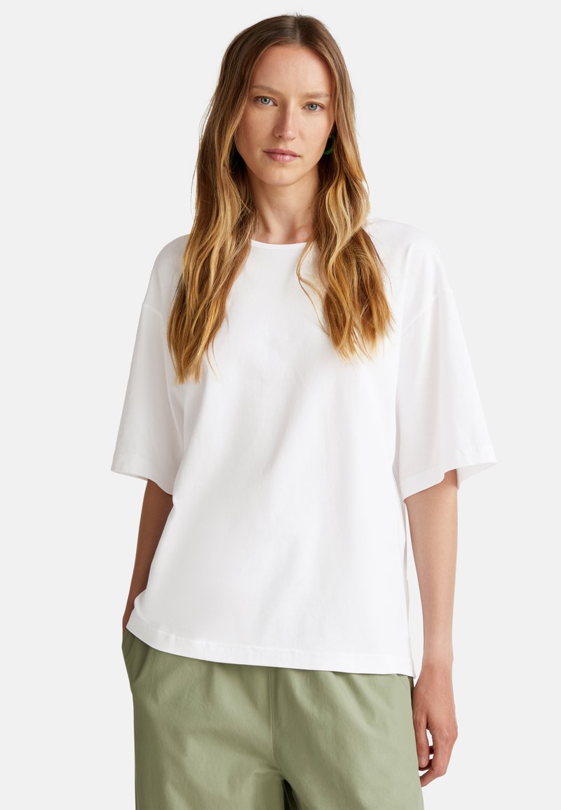 Bílý oversized tričko z jemné látky s kulatým výstřihem a krátkými rukávy, spárované se světle zelenými kalhotami. Jednoduchý, čistý design.
