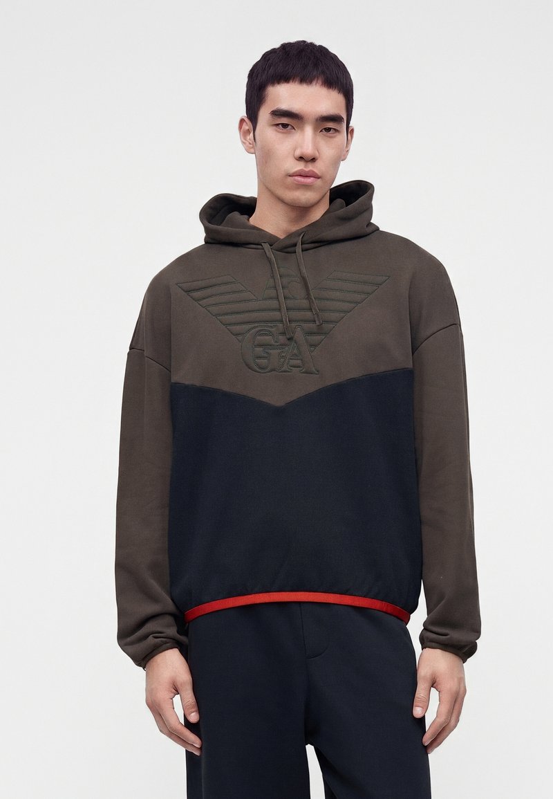 Jeune homme portant un sweat à capuche marron et noir avec un grand logo GA en relief et des ailes, debout devant un fond blanc uni.