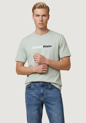 Joven con cabello rubio que lleva una camiseta verde clara de Calvin Klein y vaqueros azules, de pie con las manos juntas delante.