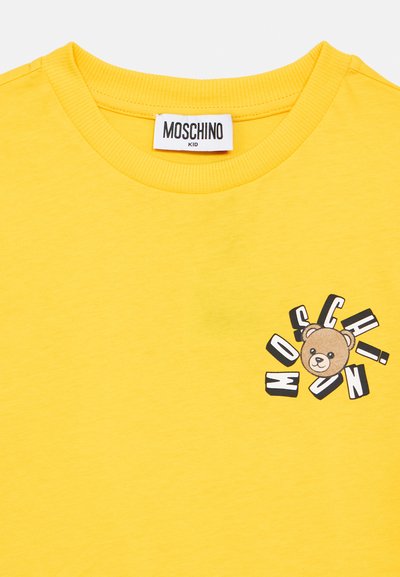 T-shirt jaune enfant avec un visage d'ours en peluche et des lettres "MOSCHINO" dispersées sur la poitrine, doté d'un col rond et d'une étiquette de marque à l'intérieur.