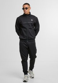 Mann in schwarzer Zip-Jacke und Hose mit weißen Turnschuhen, der vor einem schlichten hellgrauen Hintergrund steht.