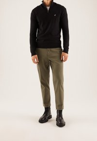 Černý svetr se zipem u krku, olivově zelené kalhoty a černé šněrovací boty. Úzký střih, ležérní design. Svetr má na hrudi malé modré logo.