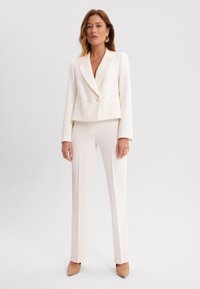 Biała, krótka marynarka z podwójnymi guzikami, w zestawie z dopasowanymi spodniami w kant. Materiał wydaje się gładki, o czystym, minimalistycznym designie.