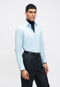 Camisa de manga longa azul clara combinada com calças pretas risca de giz e um suéter de gola alta preto, com colarinho abotoado e design ajustado.