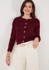 Bordeaux pletený cardigan s horizontálnymi prúžkami, s zlatými gombíkmi a okrúhlym výstrihom, skombinovaný s bielymi nohavicami a béžovým opaskom.