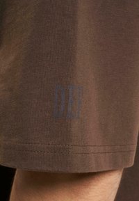 Brun bomulds t-shirt med struktureret finish; kanten viser bogstaverne "DEF" i blå. Nærbillede af stoffet og trykdetaljer.
