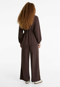 Locker sitzendes, braunes Langarmoberteil mit elastischem Bund und weit geschnittenen Hosen. Weicher Stoff mit glatter Textur und minimalen Details.