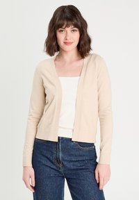 Cardigan en maille beige avec un décolleté en V et une ouverture à l'avant. Manches longues et poignets côtelés, associé à un débardeur blanc et un jean bleu marine.